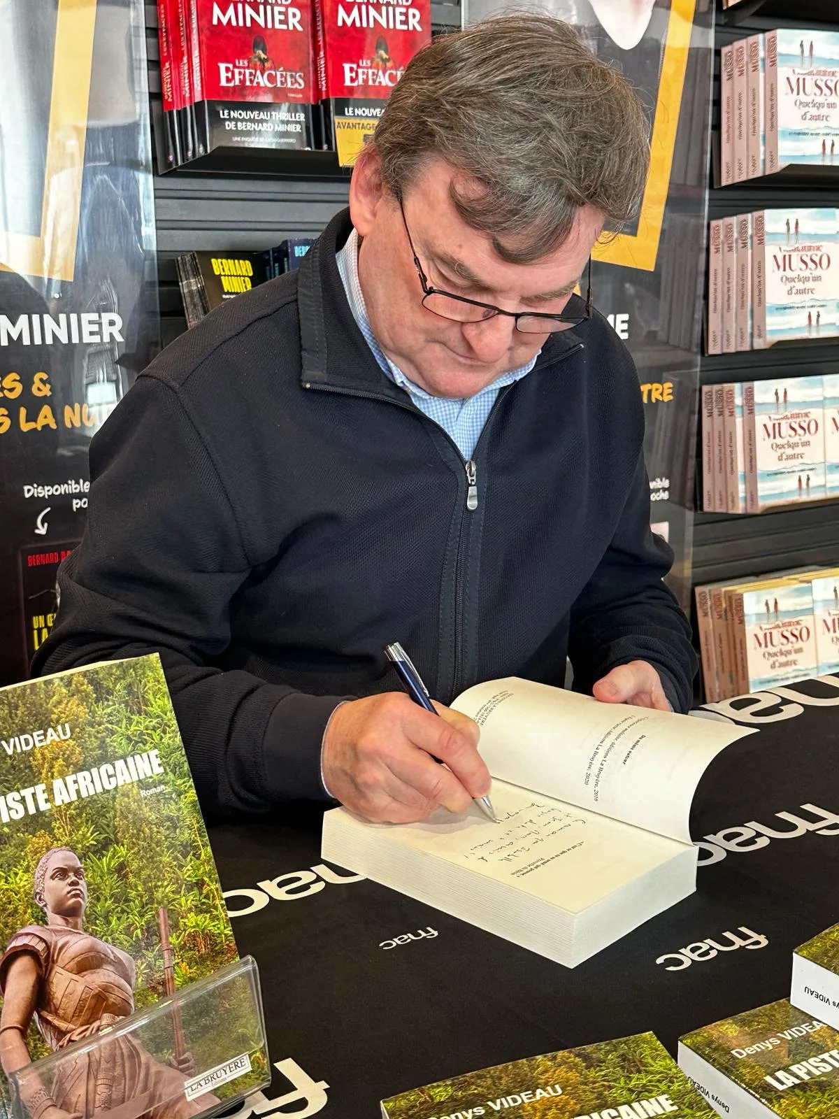 Denys Videau signant un exemplaire de La Piste africaine à la table Fnac, devant les rayons du magasin