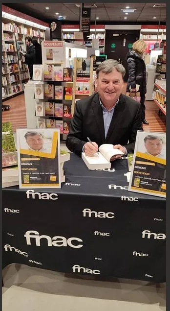 Denys Videau dédicaçant un livre à la table Fnac, entre deux affichettes annonçant la séance